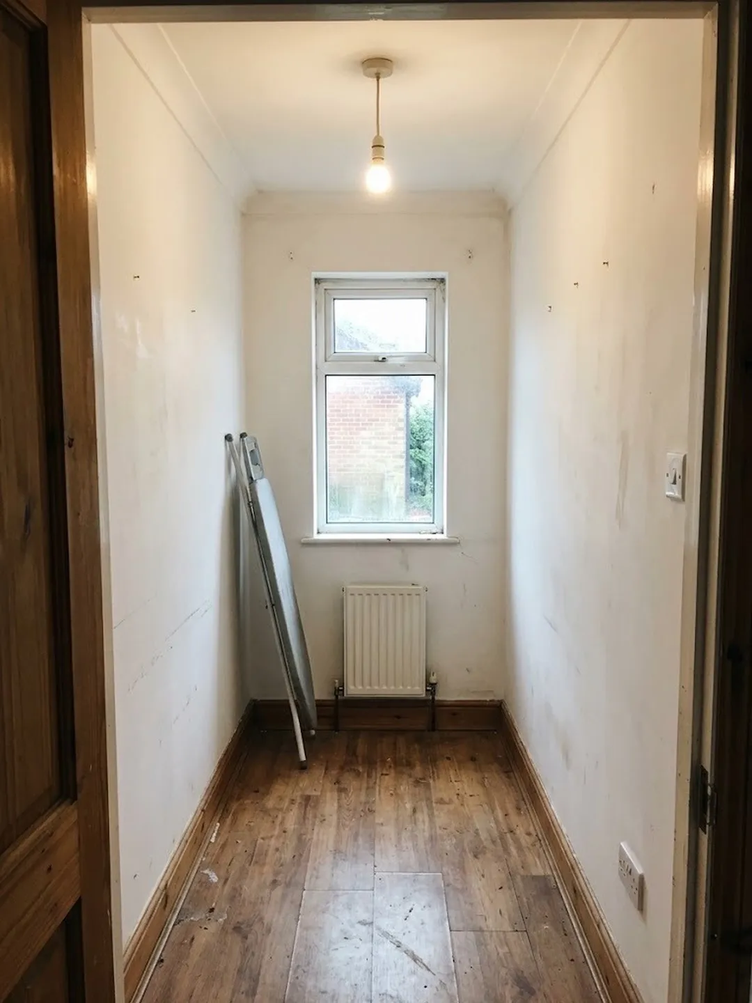 An empty narrow spare room with scuffed white walls, a bare bulb, a small window above a radiator and an ironing board leaning on the left wall
