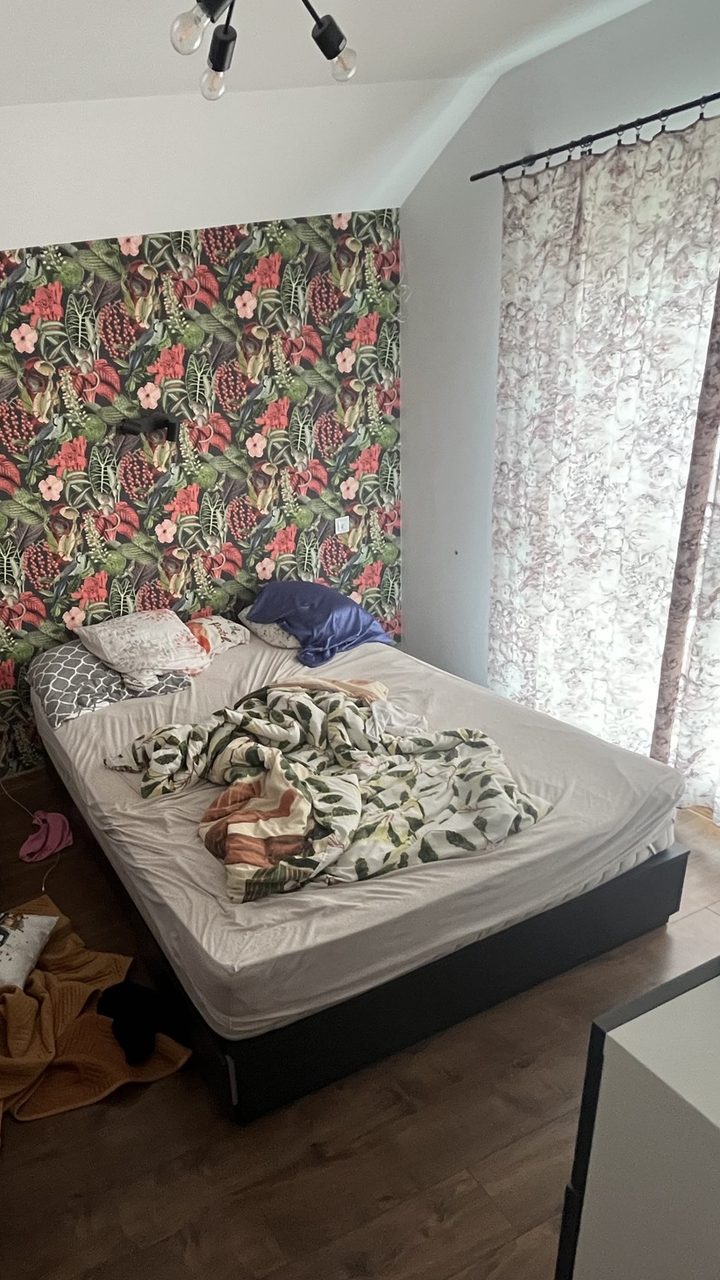 A bedroom with a bold tropical floral wallpaper wall, an unmade bed and a blanket on the floor