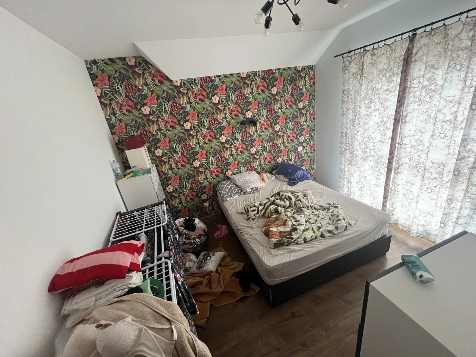 A cluttered attic bedroom with a sloped ceiling, a tropical floral wallpaper wall, patterned curtains, a black multi-arm ceiling light, an unmade bed on a black frame, a drying rack hung with laundry and clothes on the floor