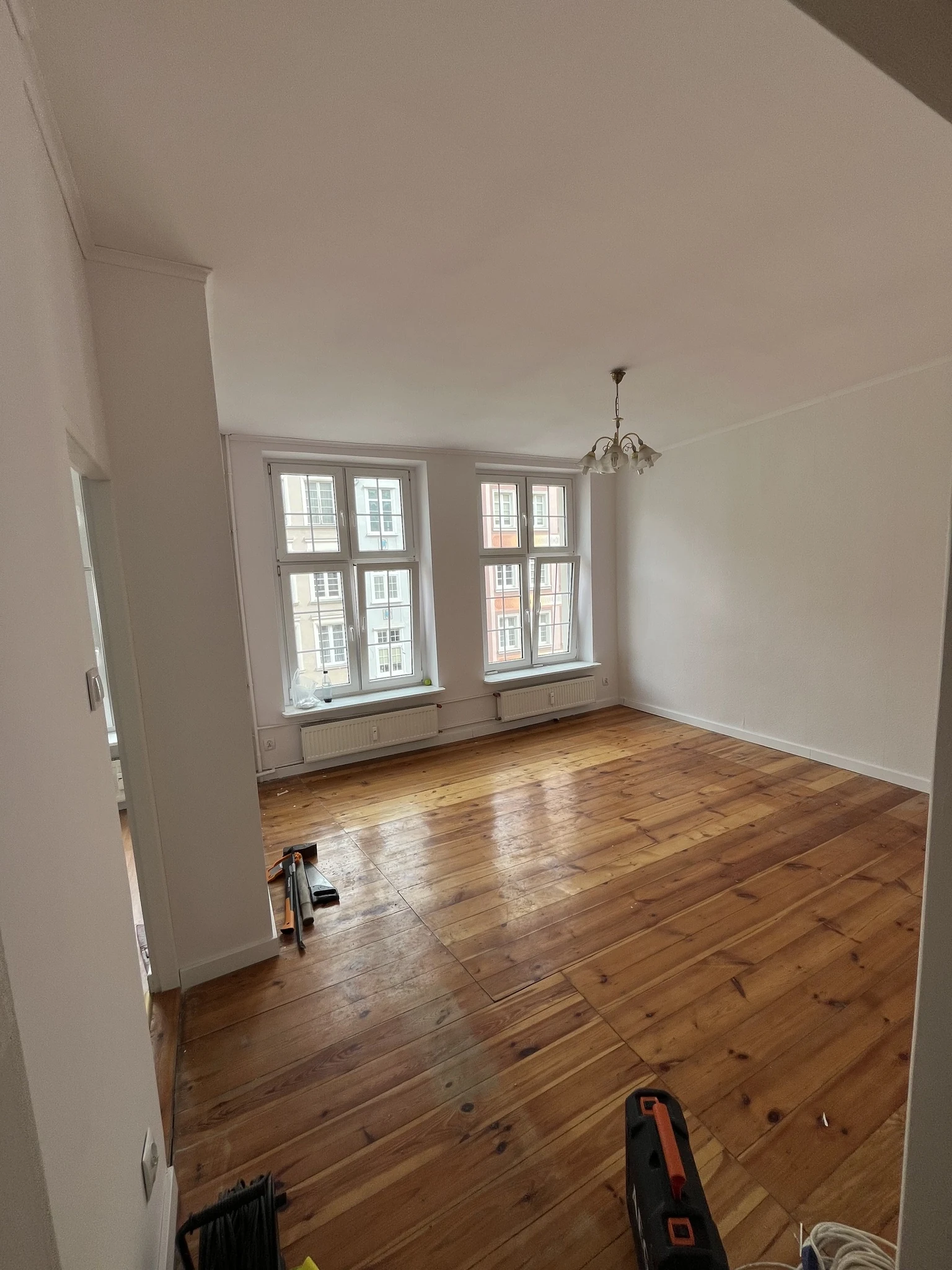 An empty, half-renovated living room with orange pine floorboards, white walls, two tall windows over radiators, a brass chandelier and tools on the floor